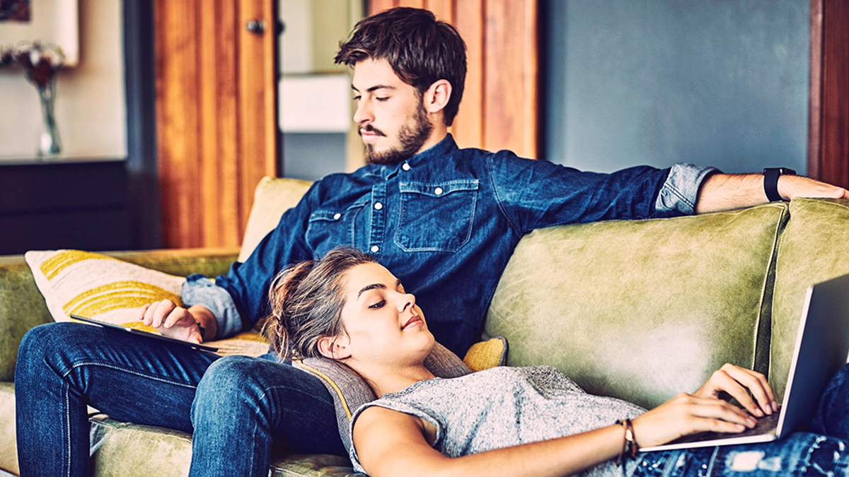 Young couple using digital tablet and laptop on sofa. Young woman with head resting on boyfriend's lap. Bloggers using technologies at home. Hipsters are in trendy casuals.