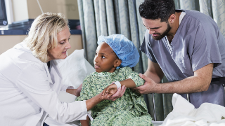 A 6 year old African-American girl sitting on a hospital gurney, with a doctor and nurse tending to her.  She is wearing a hospital gown and surgical cap. A nurse in scrubs, a young Middle Eastern man in his 20s, is standing on one side of her, and a female doctor, a mature woman in her 40s, is standing on the other side.