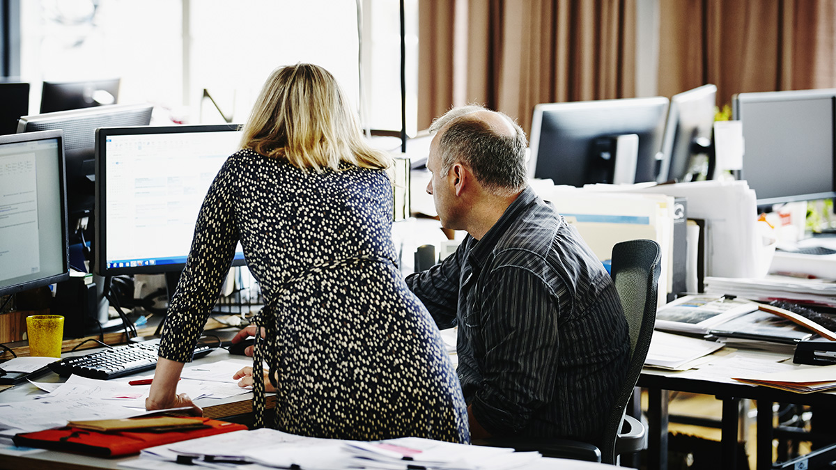 Businesswoman leaning on desk in office discussing project on computer with coworker