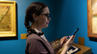 Modern museum exhibition. Side view of young Caucasian woman uses smartphone. In background are arts. Concept of visiting art gallery.
